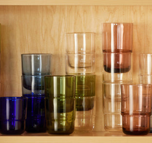 A wooden shelf displays stacked drinking glasses in various colors, including blue, green, clear, and pink. The glasses are neatly arranged in groups according to their color.