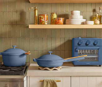 A kitchen with green tile backsplash, wooden shelves holding dishes and jars, blue Dutch oven and skillet on the counter, and a blue countertop oven beside them. A striped dish towel hangs from the cabinet handle.