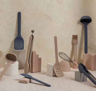 Various modern kitchen utensils in neutral and navy tones are artfully arranged upright and leaning on geometric blocks of different shapes and sizes against a beige textured background.